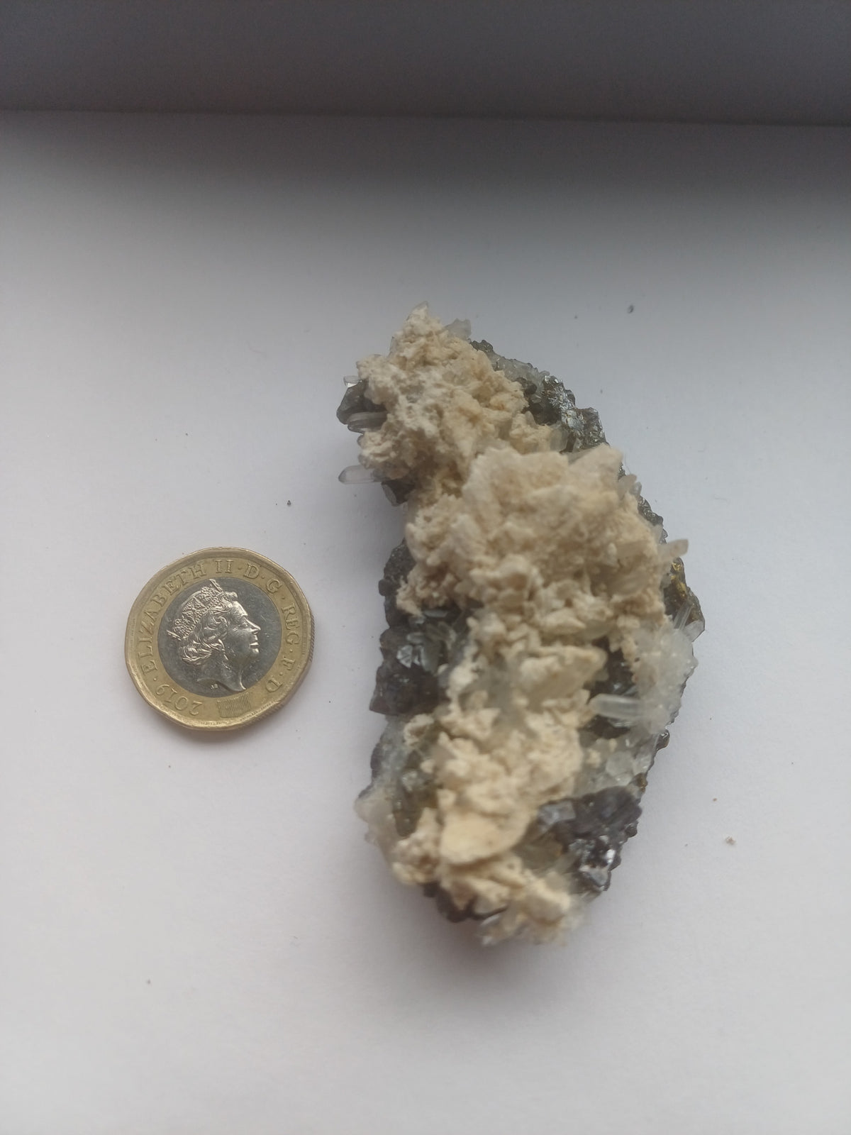 Mixed mineral specimen with dark sphalerite-like crystals, clear quartz points and pale matrix beside a £1 coin for scale